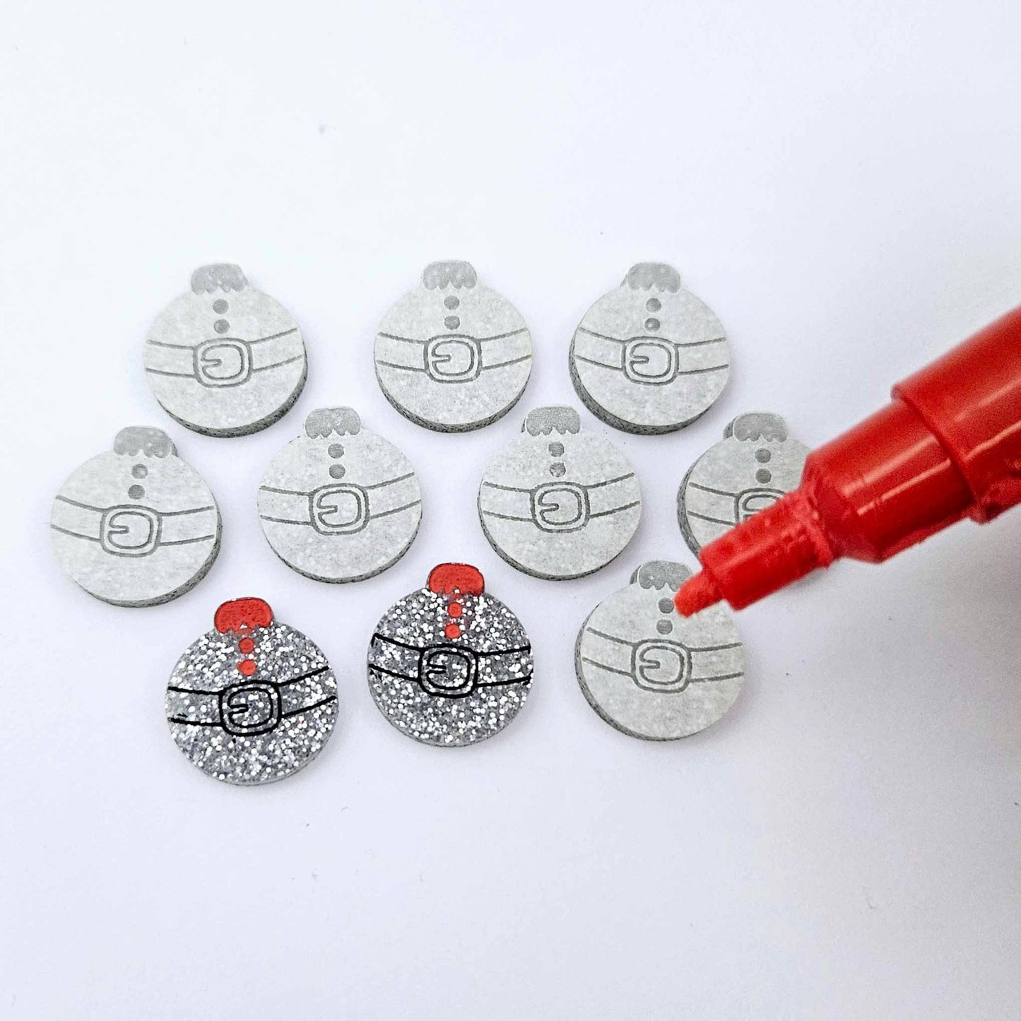 One pair of silver glitter Santa bauble stud blanks paint filled black and red alongside multiple unpainted blanks with protective paper attached, with an uncapped Posca paint pen positioned ready to paint fill the engraved details
