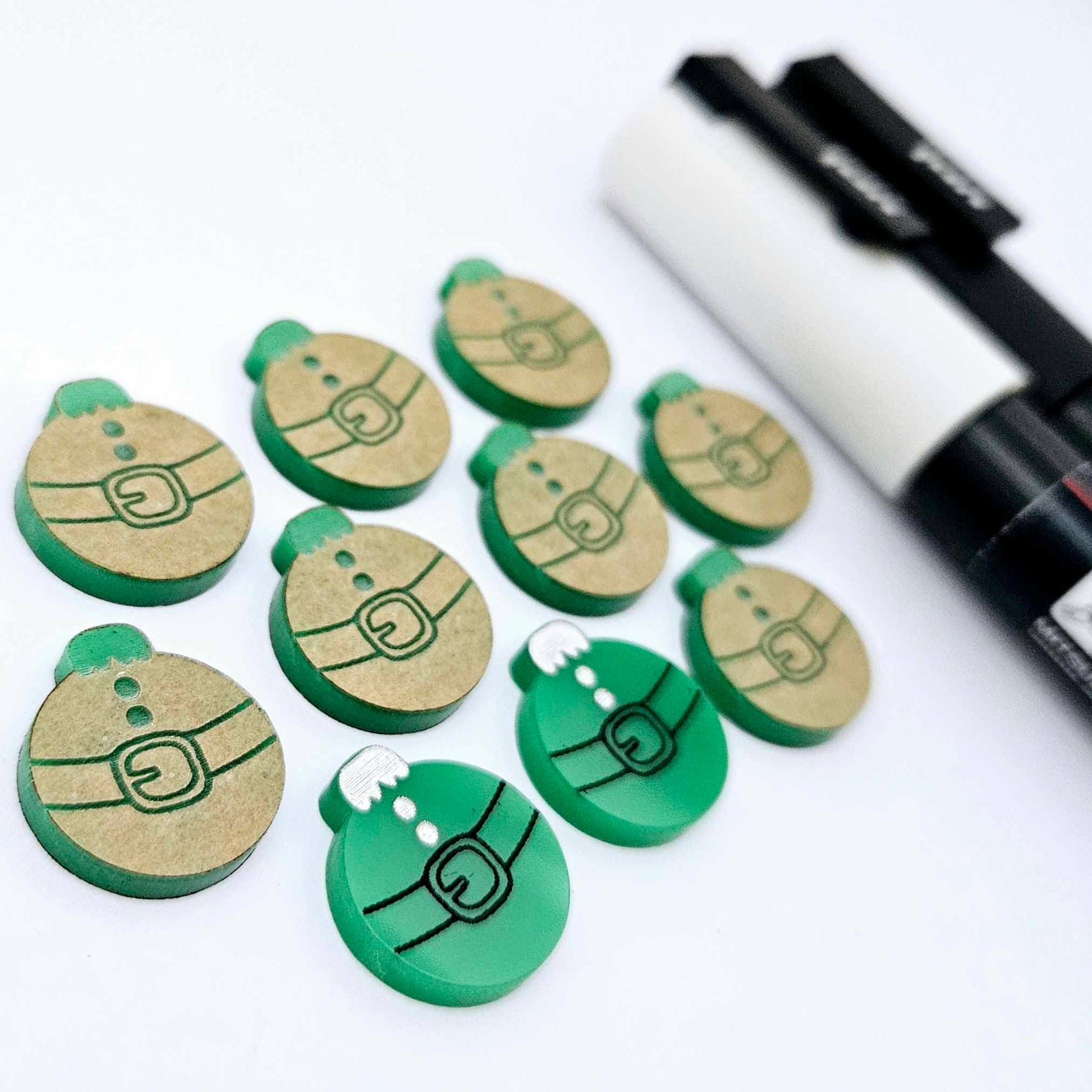 One pair of solid green Santa bauble stud blanks paint filled black and white, surrounded by multiple unpainted blanks with protective paper attached, pictured with Posca paint pens