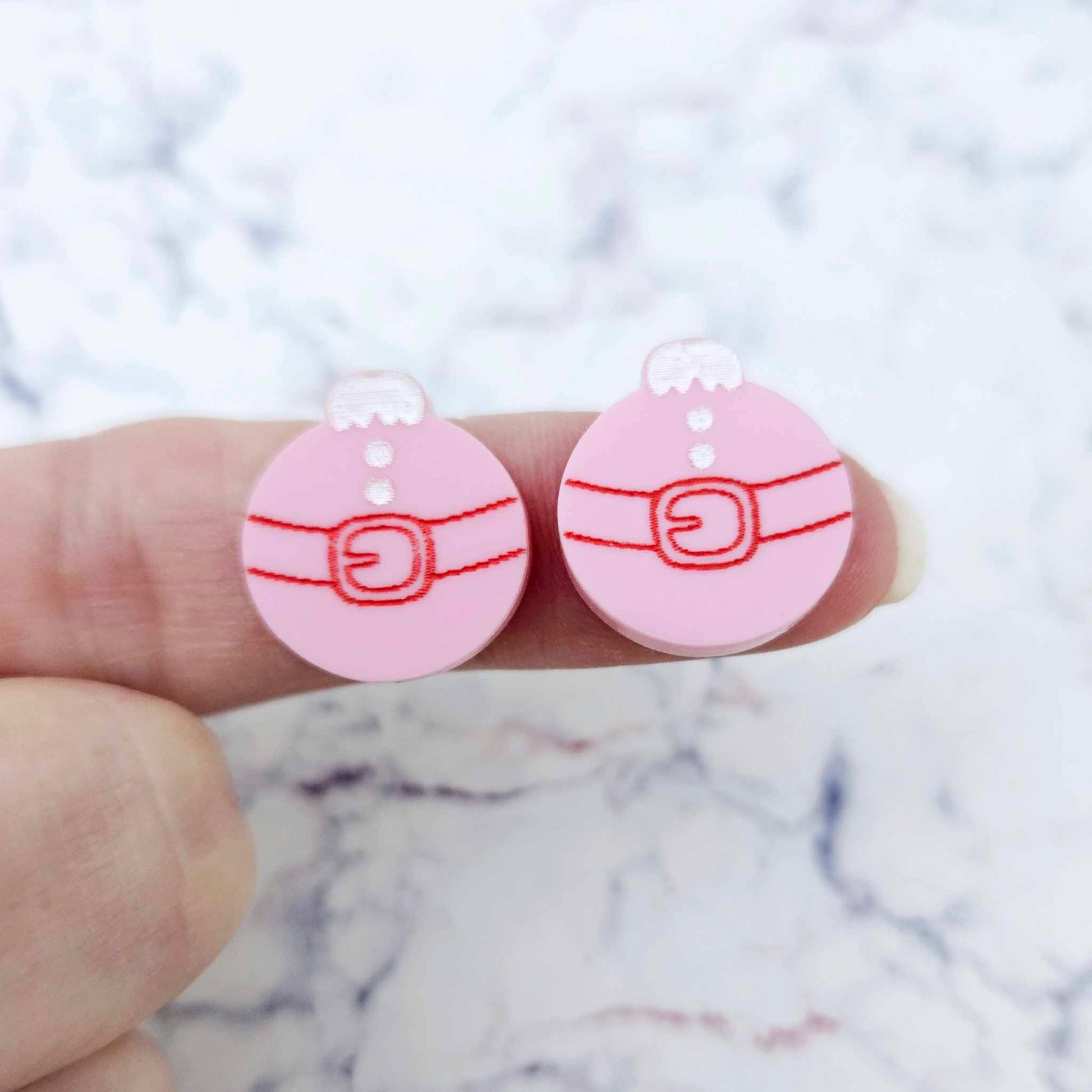 One pair of pastel pink Santa bauble stud earring blanks with engraved Santa belt and button details paint filled red and white, shown on a finger for size reference