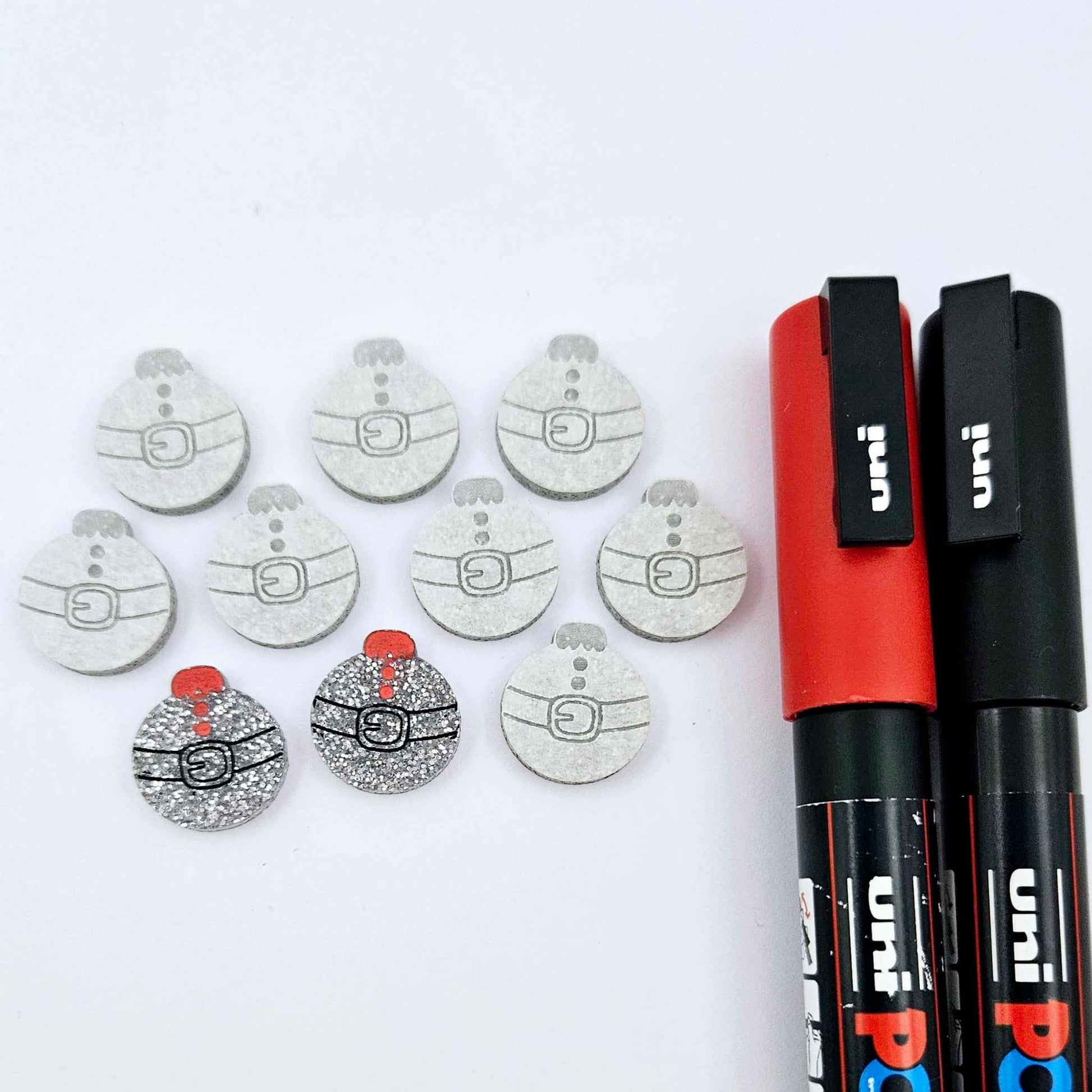 One pair of silver glitter Santa bauble stud blanks paint filled black and red, surrounded by multiple unpainted blanks with protective paper attached, pictured with Posca paint pens