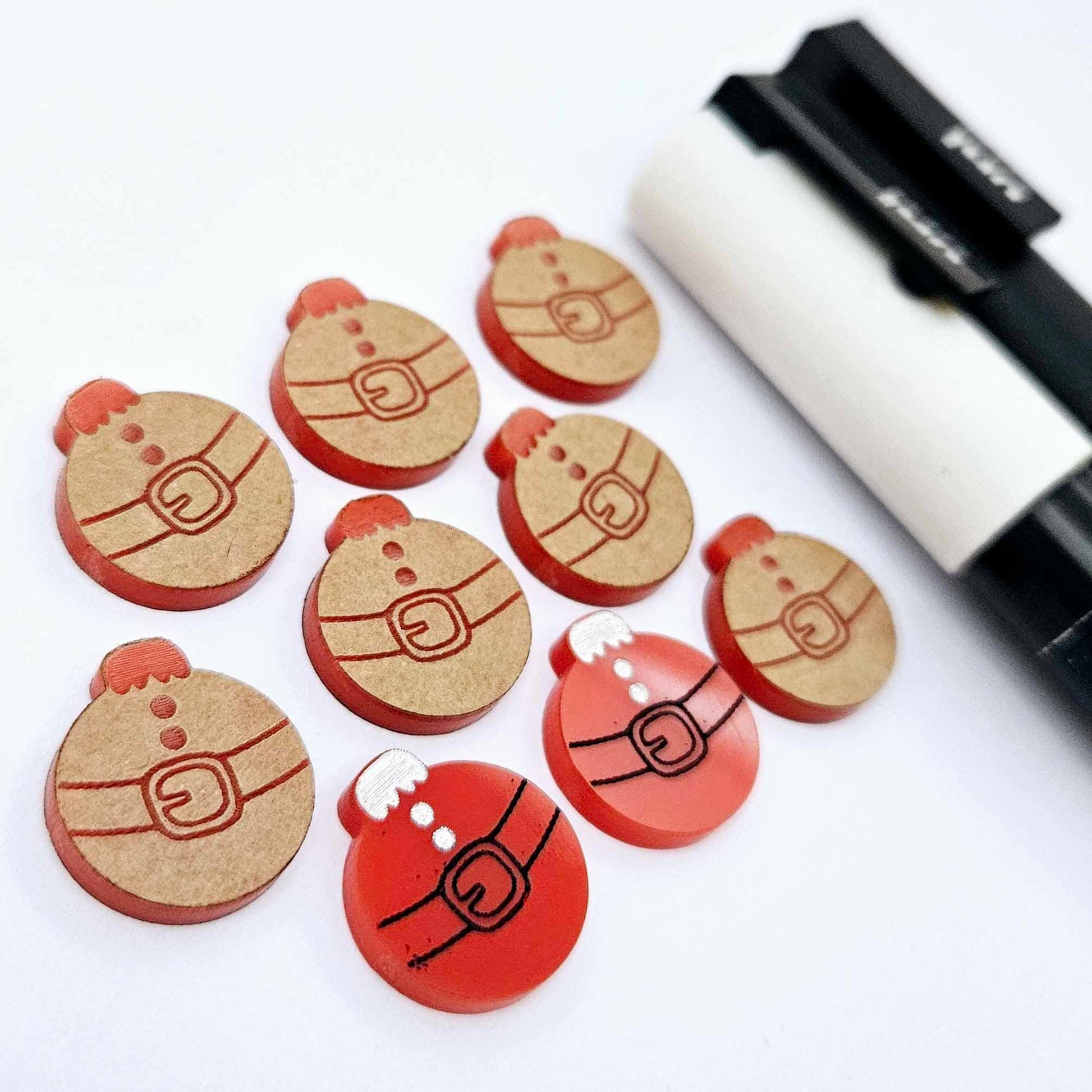 One pair of solid red Santa bauble stud blanks paint filled black and white, surrounded by multiple unpainted blanks with protective paper attached, pictured with Posca paint pens