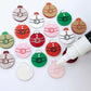 One paint-filled pair each of solid red, solid green, pastel pink, white marble and red marble Santa bauble stud blanks, with additional unpainted blanks still covered with protective paper scattered around, with an uncapped Posca paint pen positioned ready to paint fill the engraved details