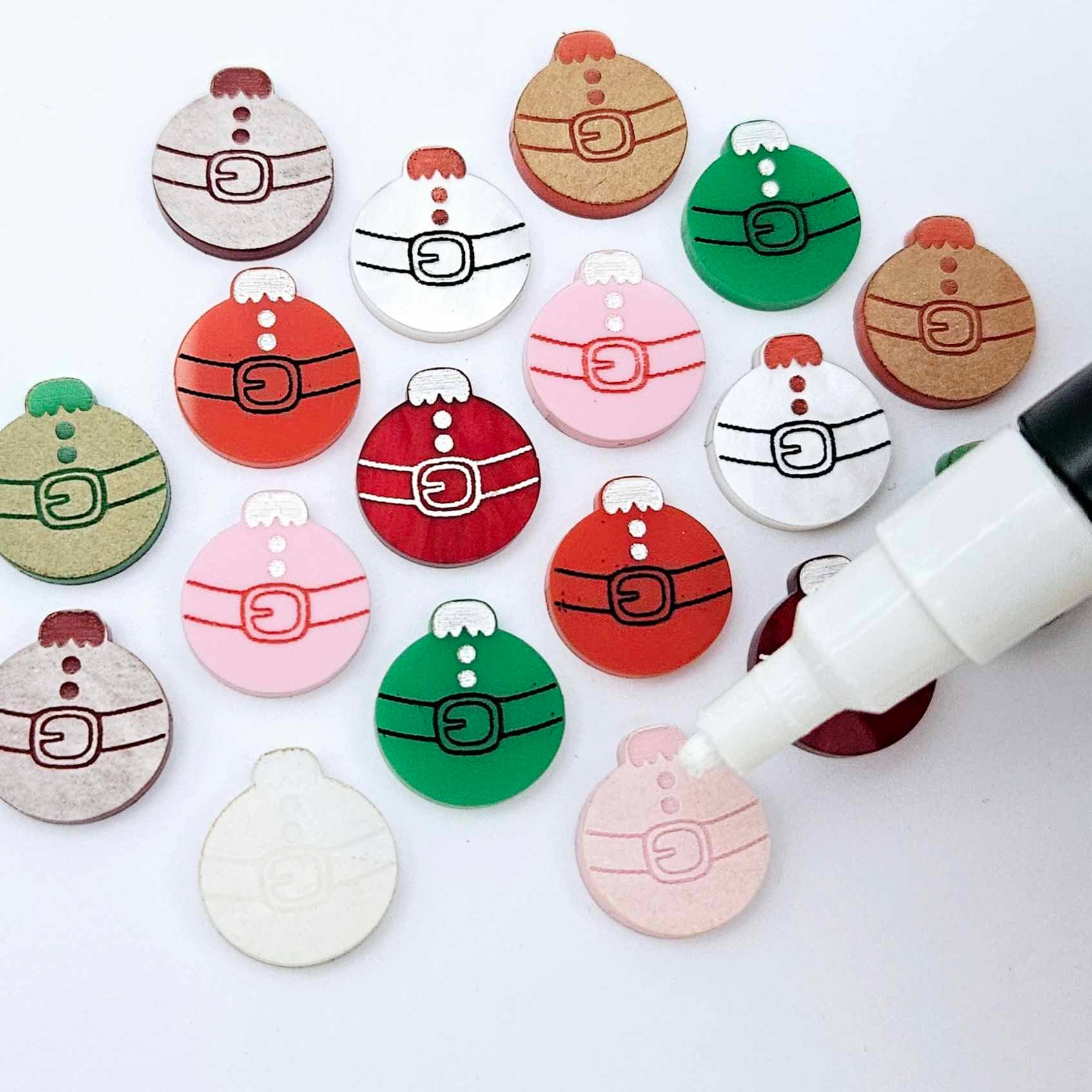 One paint-filled pair each of solid red, solid green, pastel pink, white marble and red marble Santa bauble stud blanks, with additional unpainted blanks still covered with protective paper scattered around, with an uncapped Posca paint pen positioned ready to paint fill the engraved details