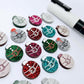 One paint-filled pair each of red glitter, hot pink glitter, teal glitter, green glitter and silver glitter Santa bauble stud blanks, with additional unpainted blanks still covered with protective paper scattered around, showing the paint-fill possibilities