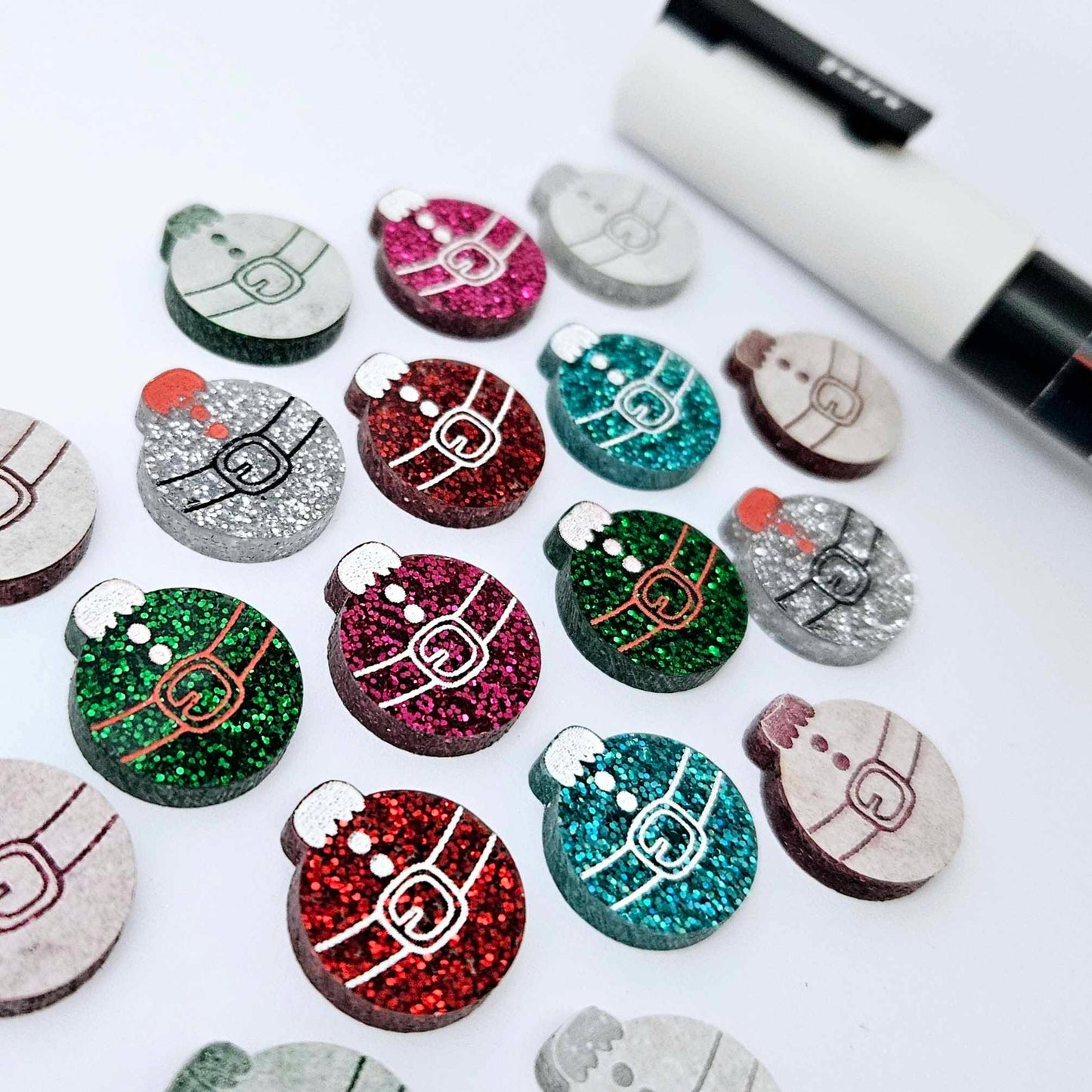 One paint-filled pair each of red glitter, hot pink glitter, teal glitter, green glitter and silver glitter Santa bauble stud blanks, with additional unpainted blanks still covered with protective paper scattered around, showing the paint-fill possibilities