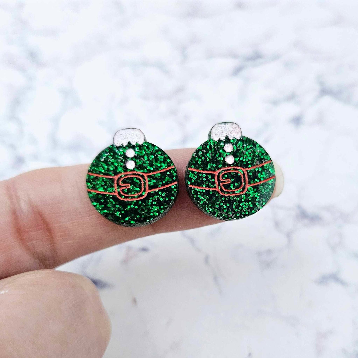One pair of green glitter Santa bauble stud earring blanks with engraved Santa belt and button details paint filled red and white, shown on a finger for size reference