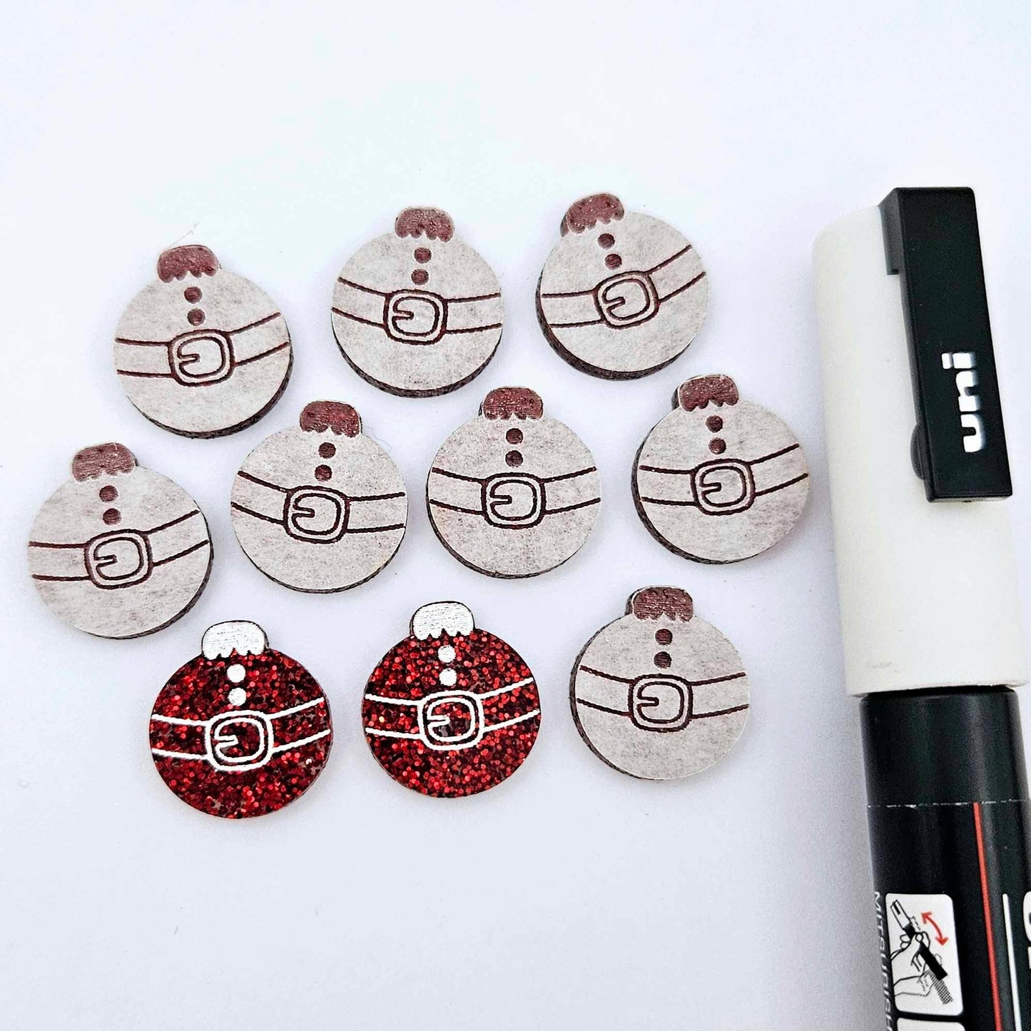 One pair of red glitter Santa bauble stud blanks paint filled white, surrounded by multiple unpainted blanks with protective paper attached, pictured with a Posca paint pen