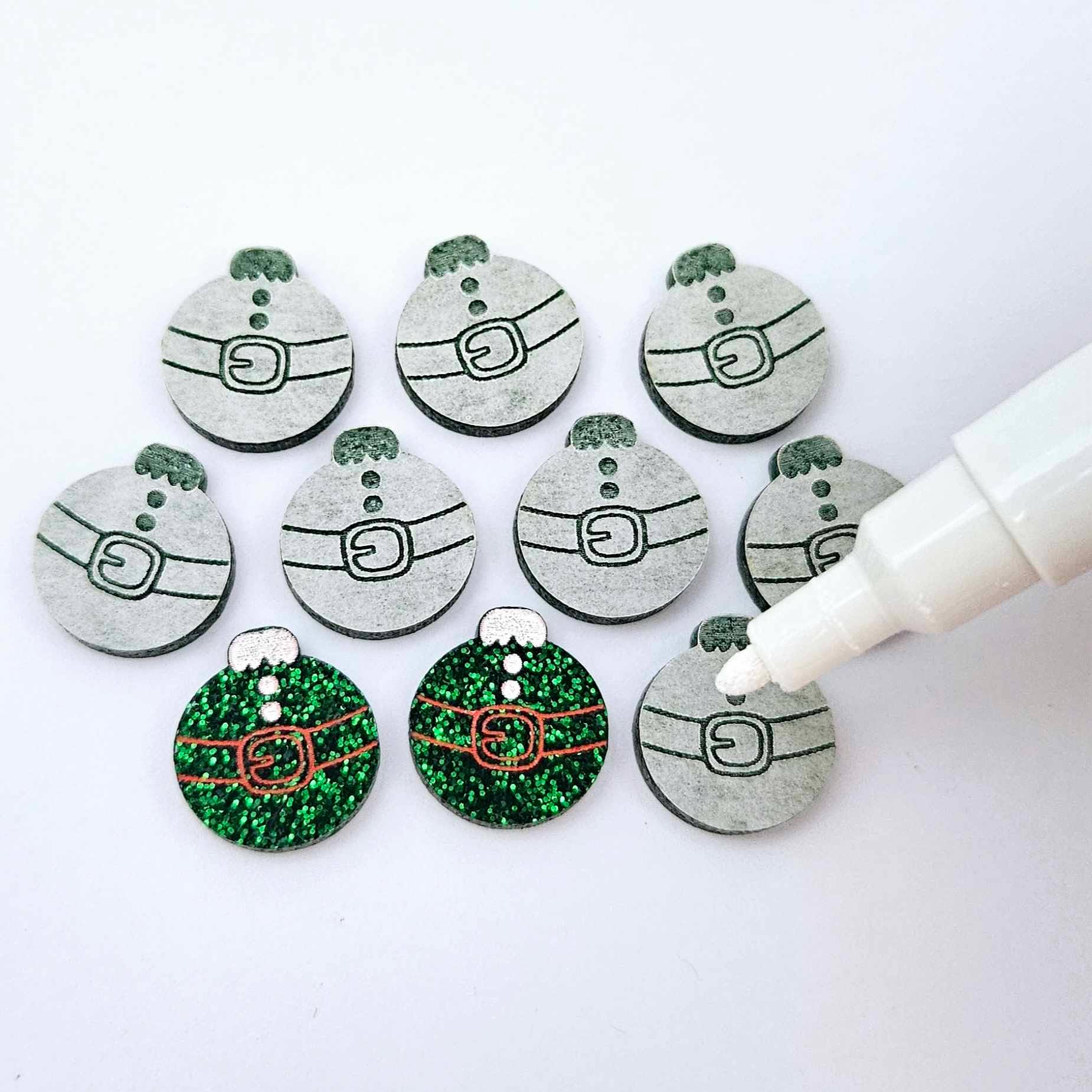 One pair of green glitter Santa bauble stud blanks paint filled red and white, surrounded by multiple unpainted blanks with protective paper attached, pictured with Posca paint pens