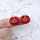 One pair of deep red marble Santa bauble stud earrings with engraved Santa belt and button details paint filled white, shown on a finger for size reference