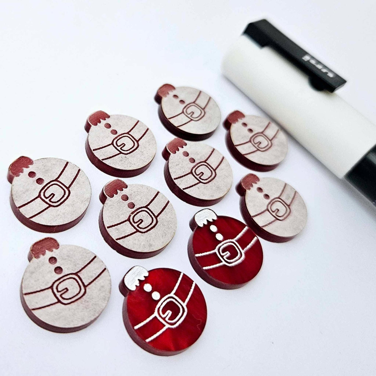 One pair of deep red marble Santa bauble stud blanks paint filled white, surrounded by multiple unpainted blanks with protective paper attached, pictured with a Posca paint pen