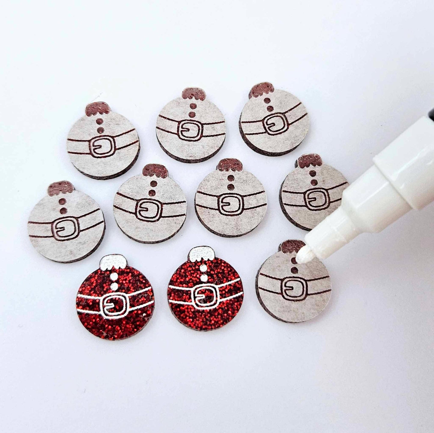 One pair of red glitter Santa bauble stud blanks paint filled white alongside multiple unpainted blanks with protective paper attached, with an uncapped Posca paint pen positioned ready to paint fill the engraved details