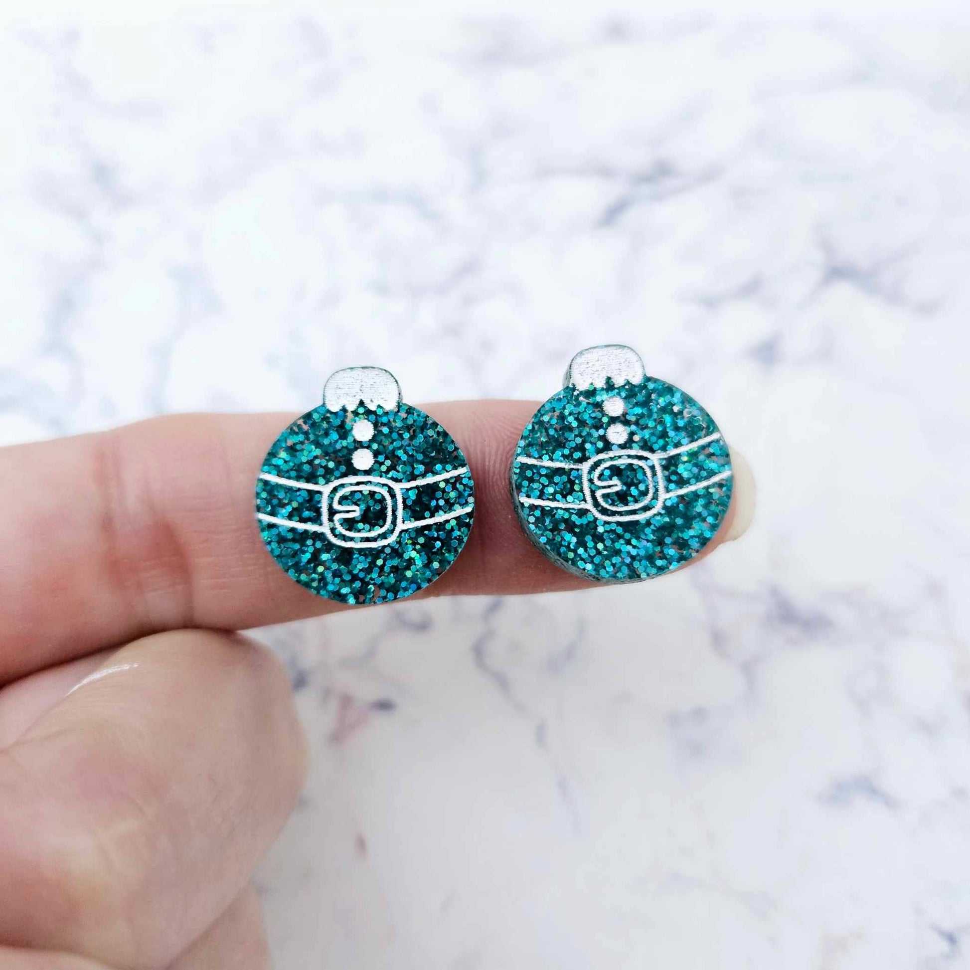 One pair of teal glitter Santa bauble stud earring blanks with engraved Santa belt and button details paint filled white, shown on a finger for size reference