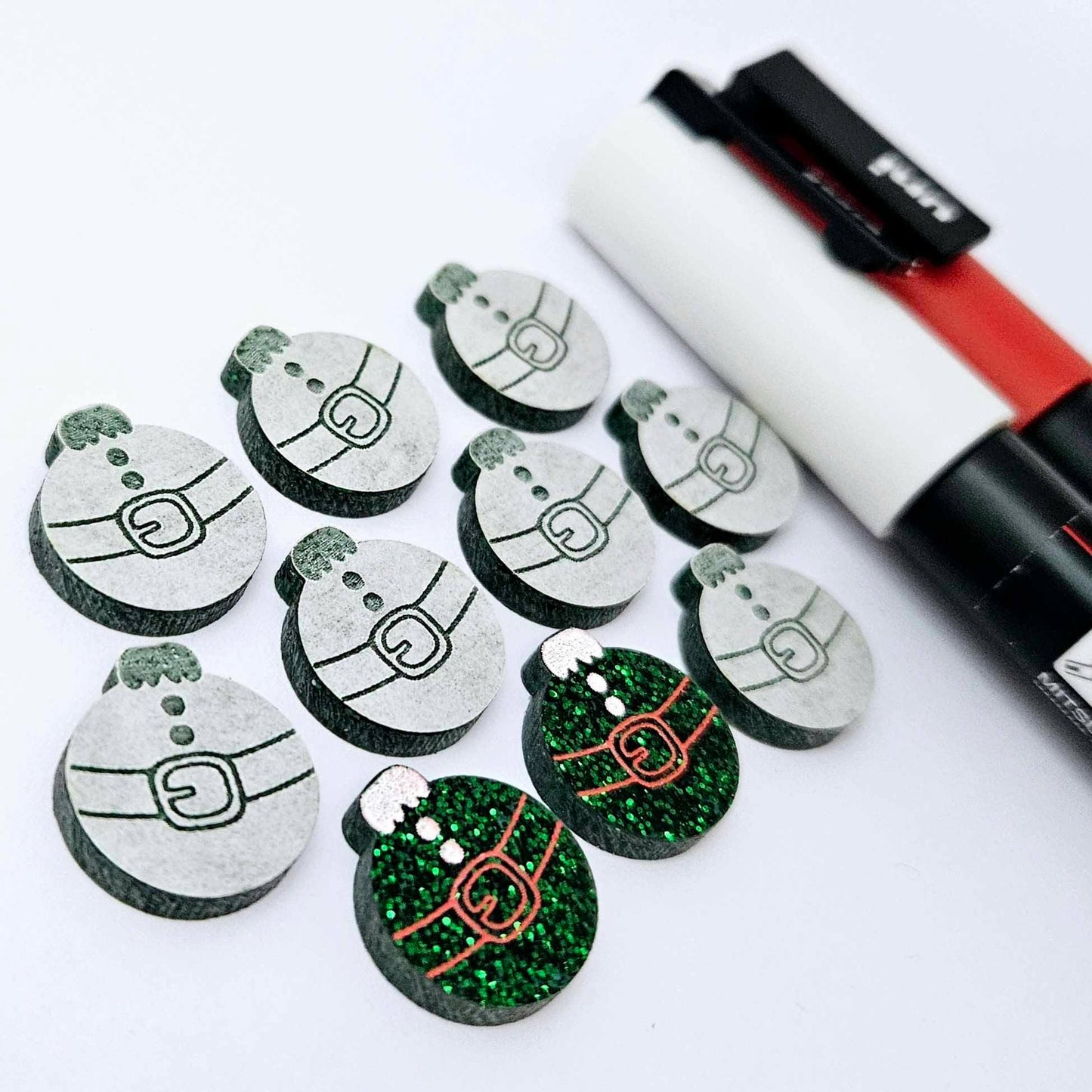 One pair of green glitter Santa bauble stud blanks paint filled red and white, surrounded by multiple unpainted blanks with protective paper attached, pictured with Posca paint pens
