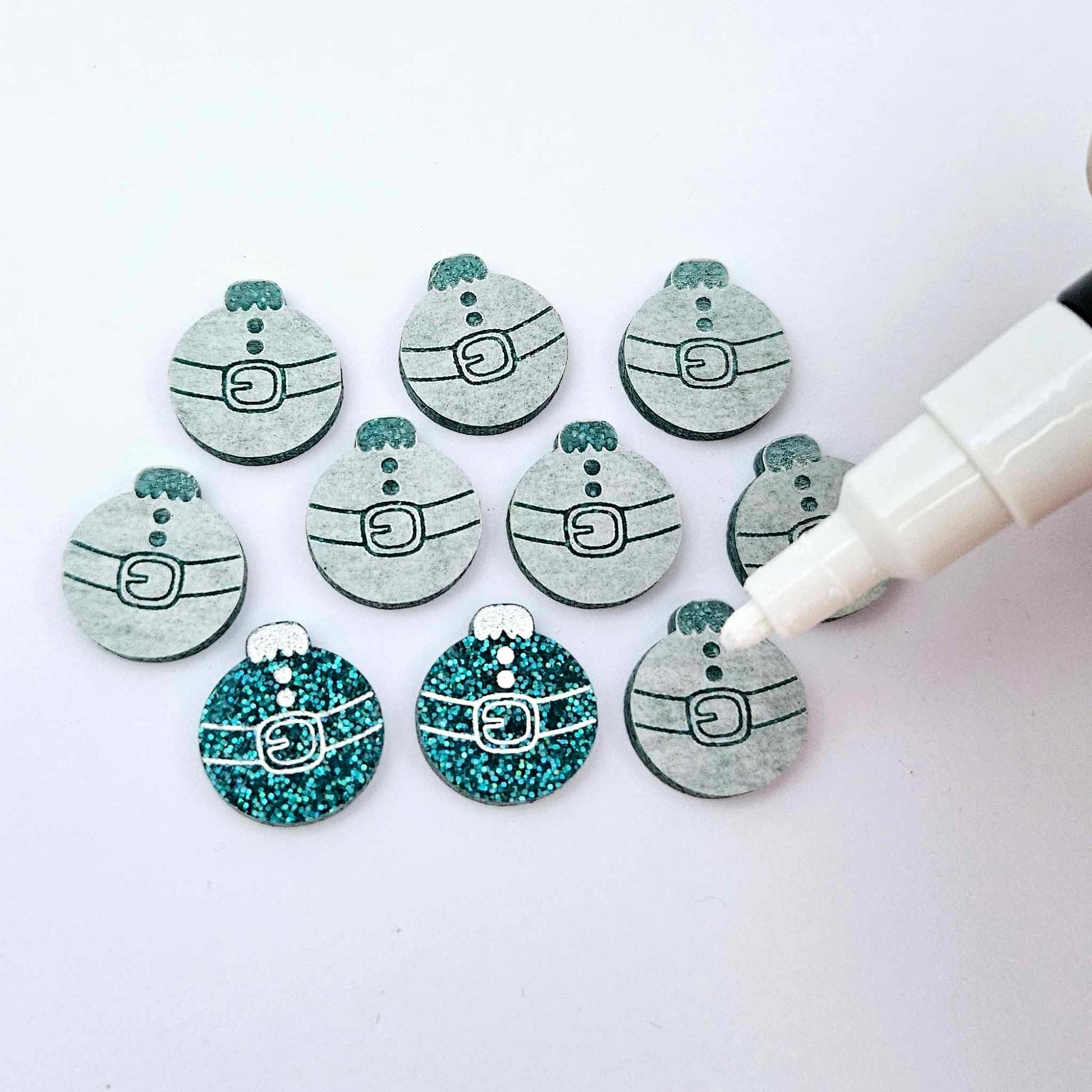One pair of teal glitter Santa bauble stud blanks paint filled white alongside multiple unpainted blanks with protective paper attached, with an uncapped Posca paint pen positioned ready to paint fill the engraved details