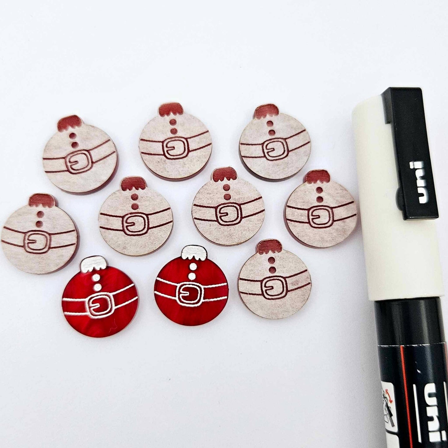 One pair of deep red marble Santa bauble stud blanks paint filled white, surrounded by multiple unpainted blanks with protective paper attached, pictured with a Posca paint pen
