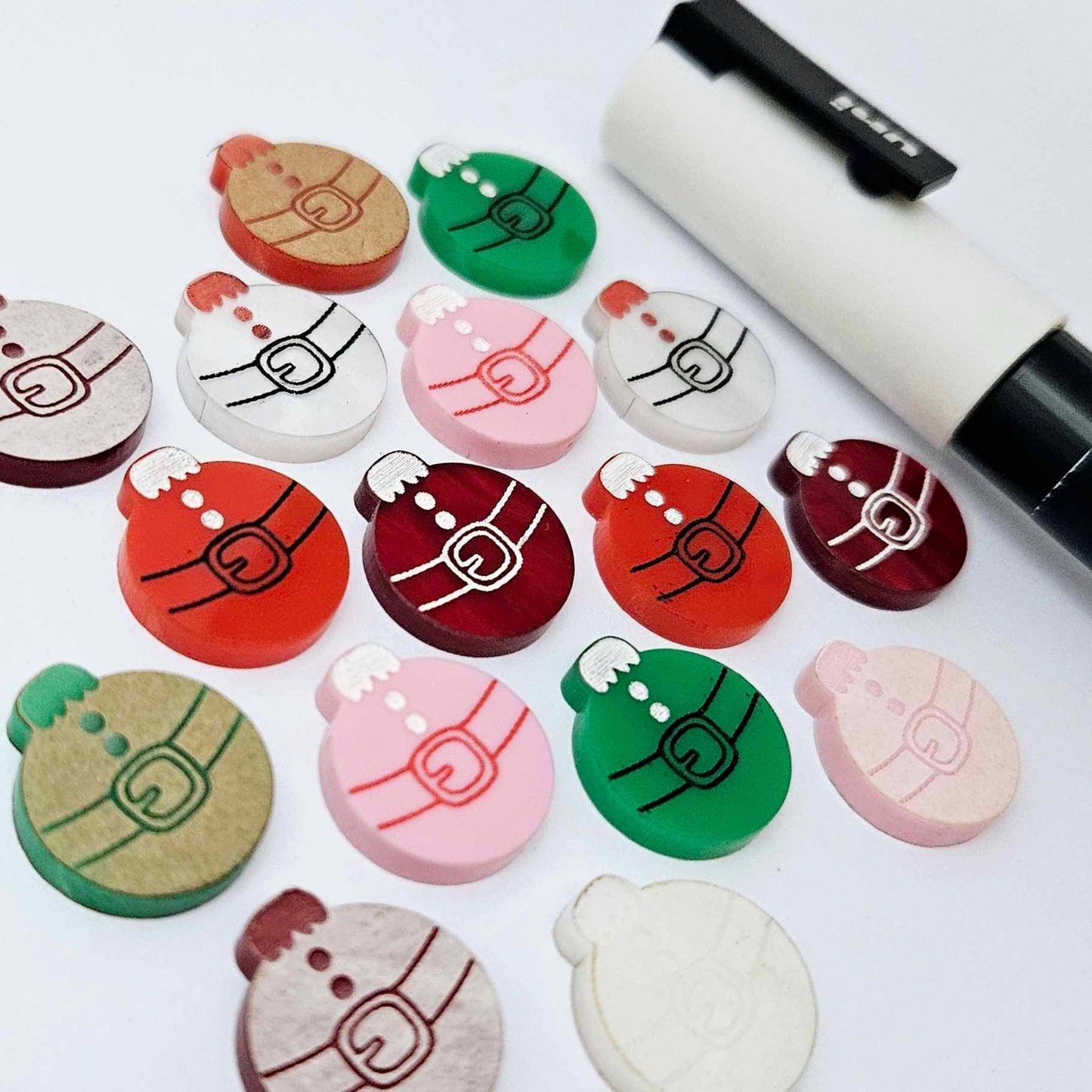One paint-filled pair each of solid red, solid green, pastel pink, white marble and red marble Santa bauble stud blanks, with additional unpainted blanks still covered with protective paper scattered around, showing the paint-fill possibilities