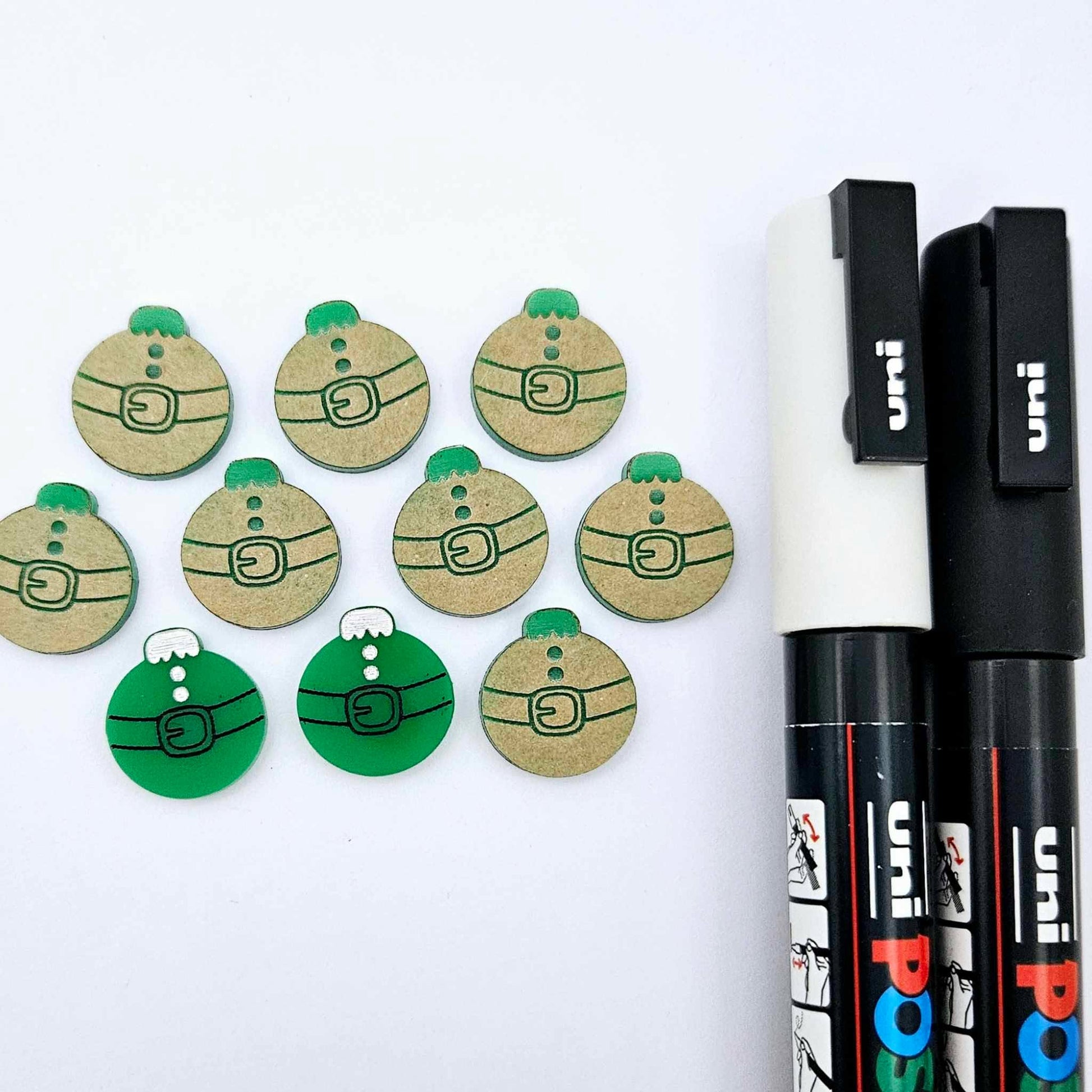One pair of solid green Santa bauble stud blanks paint filled black and white, surrounded by multiple unpainted blanks with protective paper attached, pictured with Posca paint pens