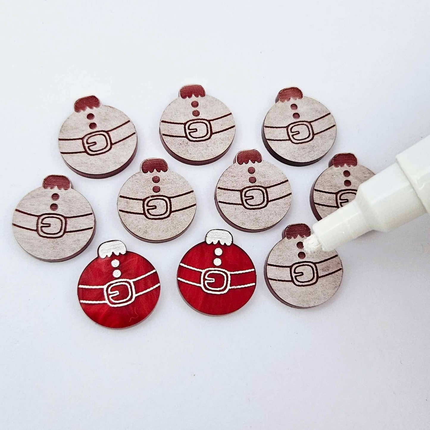 One pair of deep red marble Santa bauble stud blanks paint filled white alongside multiple unpainted blanks with protective paper attached, with an uncapped Posca paint pen positioned ready to paint fill the engraved details