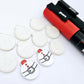 One pair of white marble Santa bauble stud blanks paint filled red and black, surrounded by multiple unpainted blanks with protective paper attached, pictured with Posca paint pens