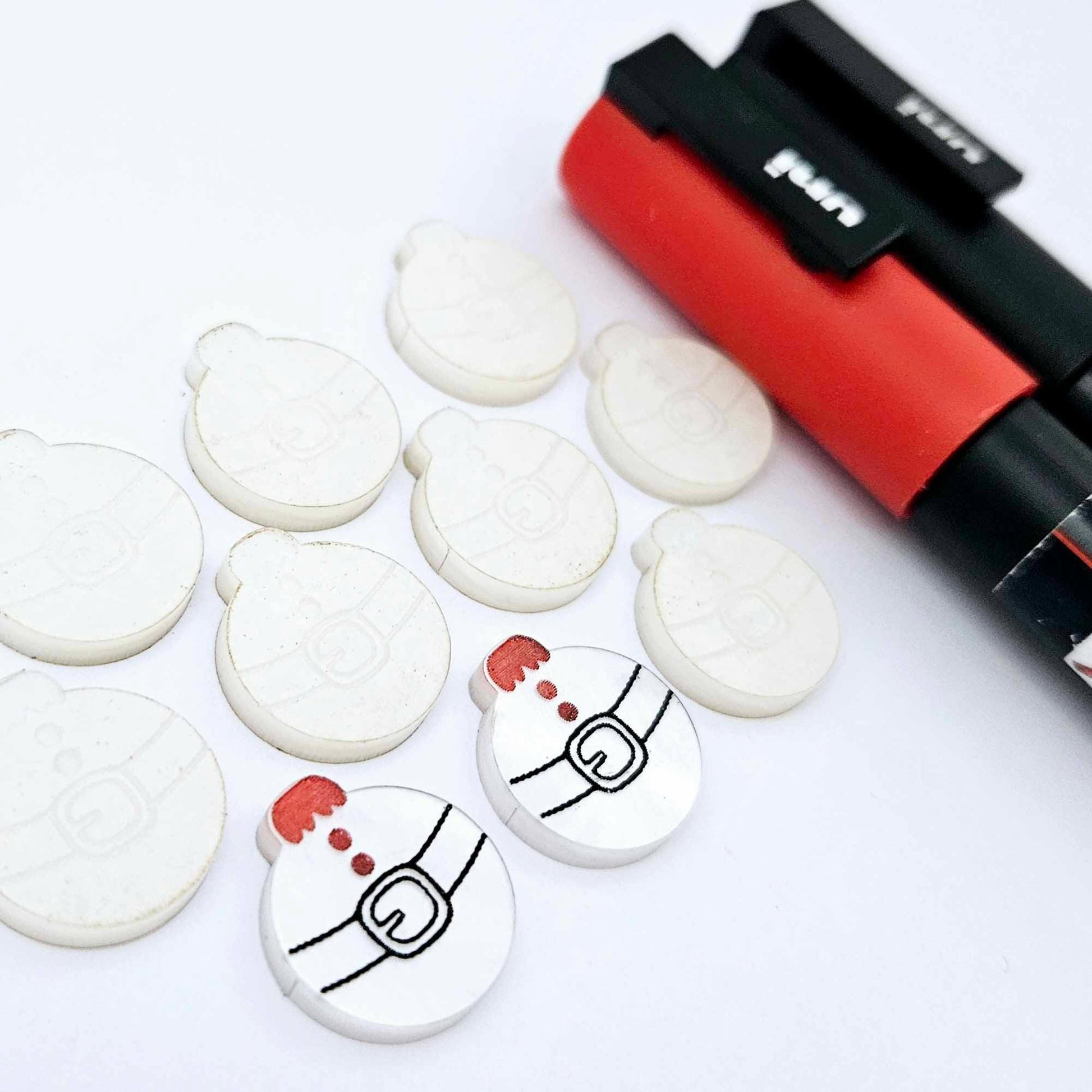 One pair of white marble Santa bauble stud blanks paint filled red and black, surrounded by multiple unpainted blanks with protective paper attached, pictured with Posca paint pens