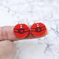 One pair of solid red Santa bauble stud earring blanks with engraved Santa belt and button details paint filled black and white, shown on a finger for size reference