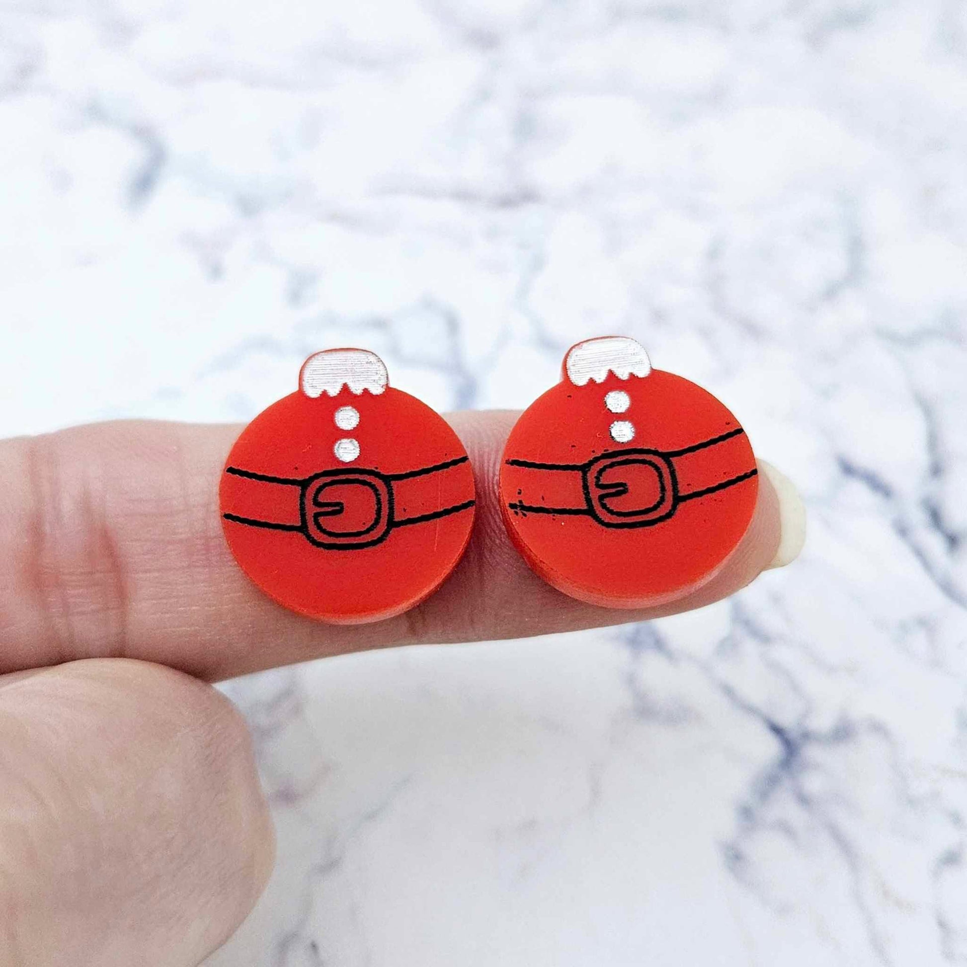 One pair of solid red Santa bauble stud earring blanks with engraved Santa belt and button details paint filled black and white, shown on a finger for size reference