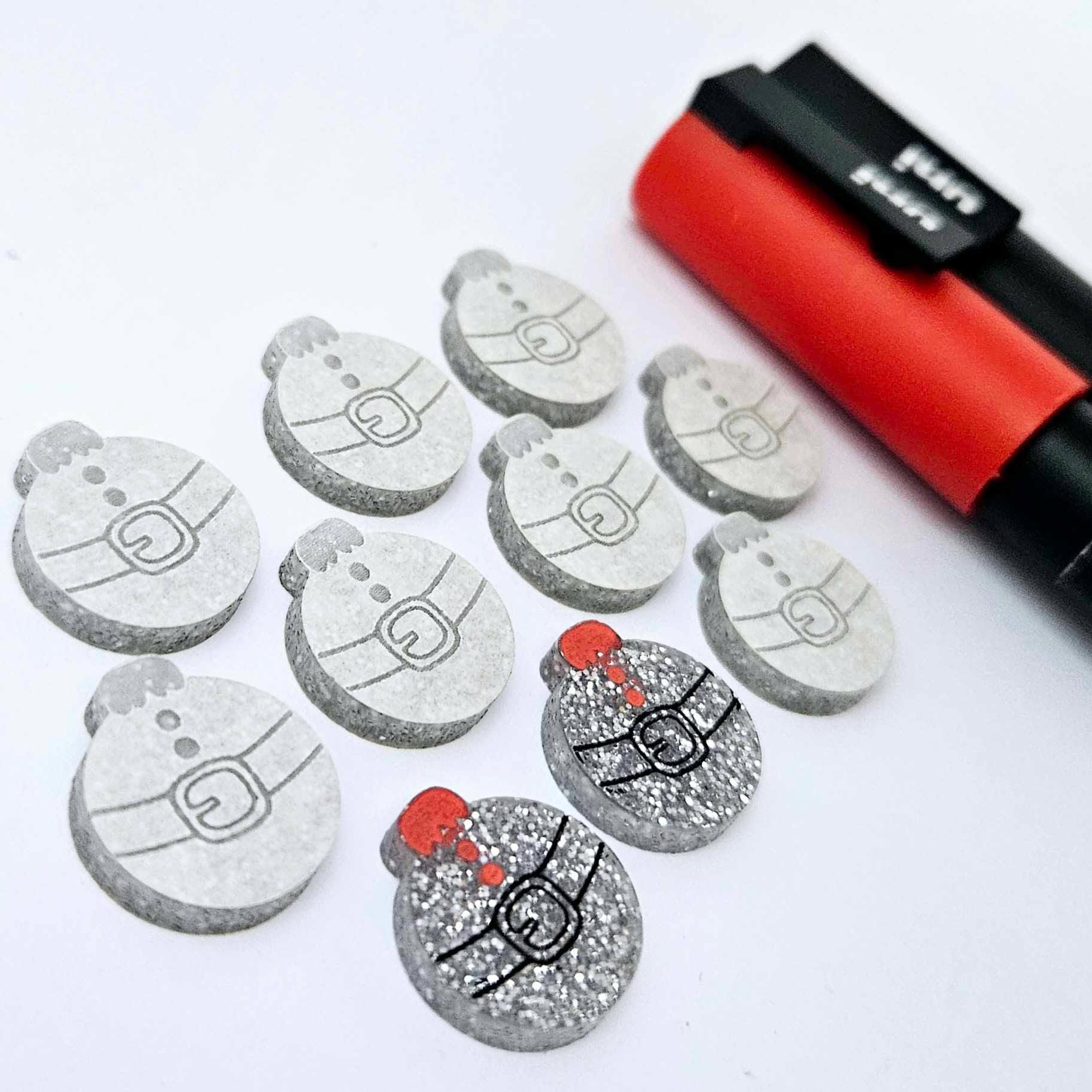 One pair of silver glitter Santa bauble stud blanks paint filled black and red, surrounded by multiple unpainted blanks with protective paper attached, pictured with Posca paint pens