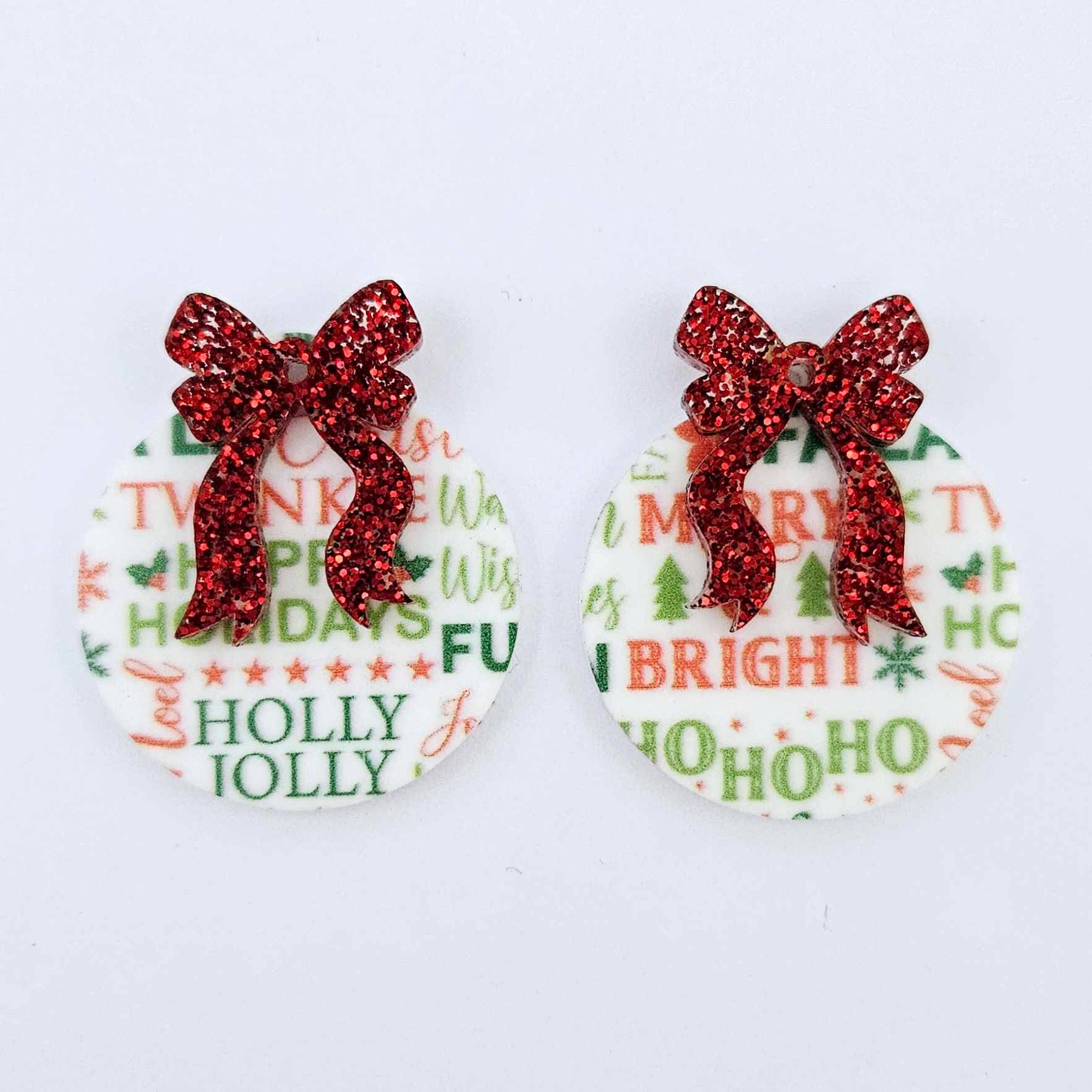 One pair of Christmas Text UV printed bauble dangle blanks with included red glitter bows laying flat.