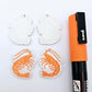 One pair of white marble acrylic engraved prawn dangle earring blanks with protective paper on alongside a paint filled pair, laying flat with an orange posca paint pen