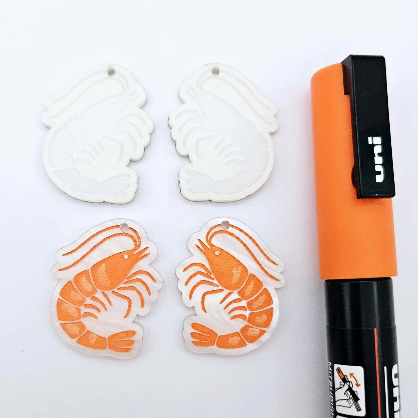 One pair of white marble acrylic engraved prawn dangle earring blanks with protective paper on alongside a paint filled pair, laying flat with an orange posca paint pen