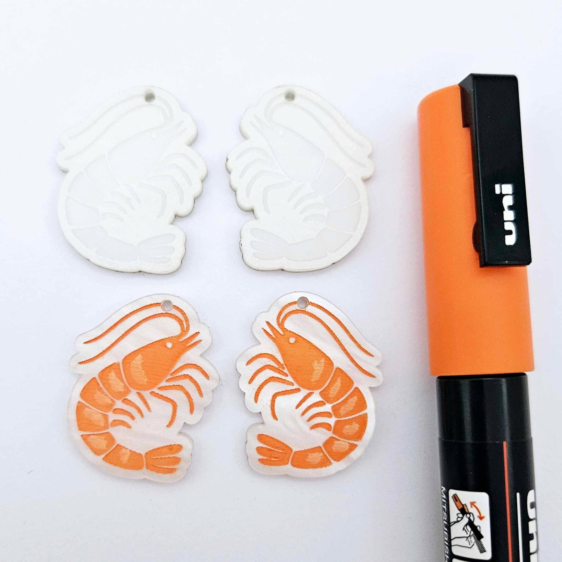 One pair of white marble acrylic engraved prawn dangle earring blanks with protective paper on alongside a paint filled pair, laying flat with an orange posca paint pen