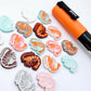 A selection of paint-filled prawn stud earring blanks in Pastel Pink, Pastel Aqua, Red Glitter, Silver Glitter and White Marble acrylic, with multiple unpainted pieces still covered with protective paper, pictured alongside an Orange Posca Paint Pen