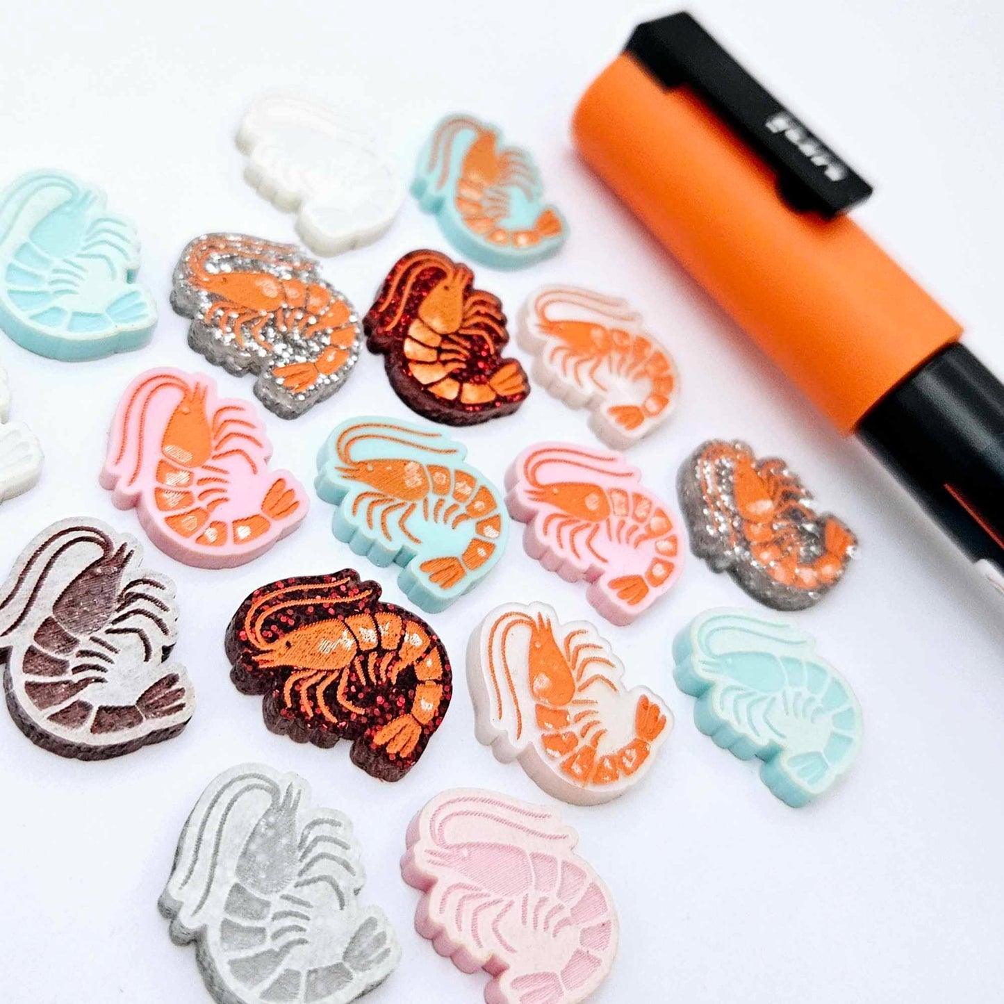 A selection of paint-filled prawn stud earring blanks in Pastel Pink, Pastel Aqua, Red Glitter, Silver Glitter and White Marble acrylic, with multiple unpainted pieces still covered with protective paper, pictured alongside an Orange Posca Paint Pen