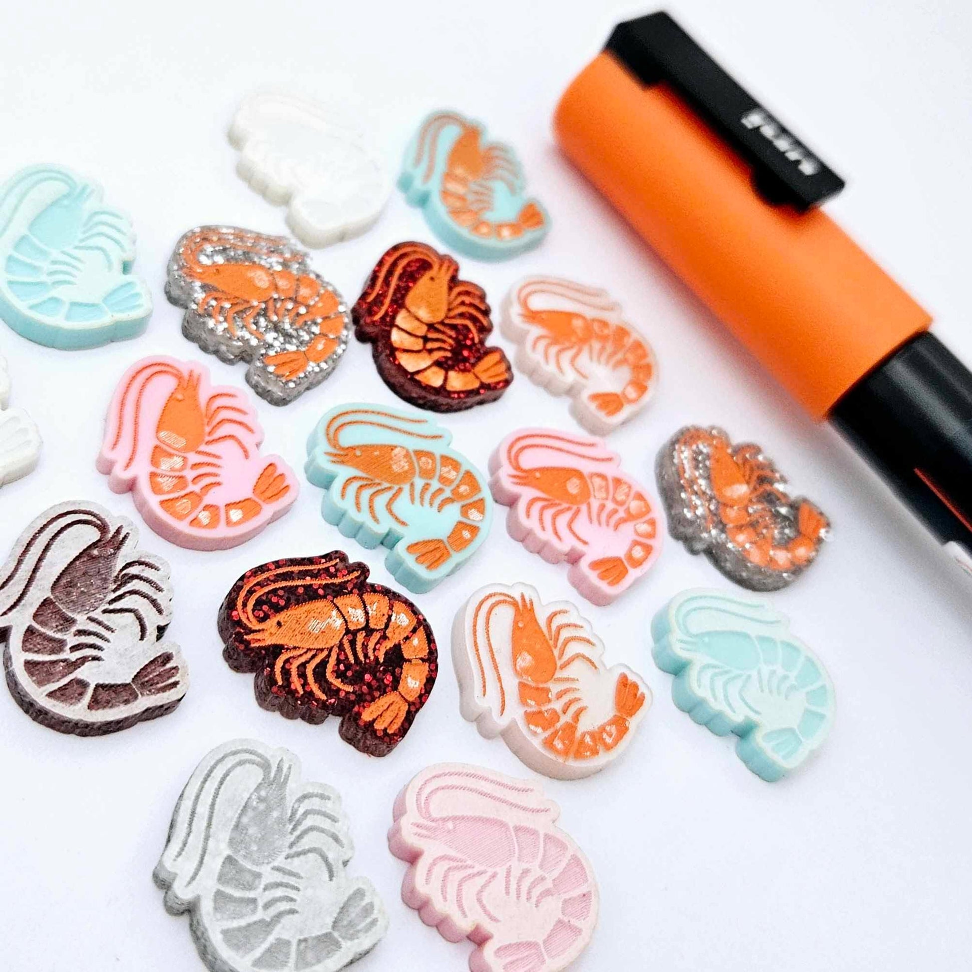 A selection of paint-filled prawn stud earring blanks in Pastel Pink, Pastel Aqua, Red Glitter, Silver Glitter and White Marble acrylic, with multiple unpainted pieces still covered with protective paper, pictured alongside an Orange Posca Paint Pen