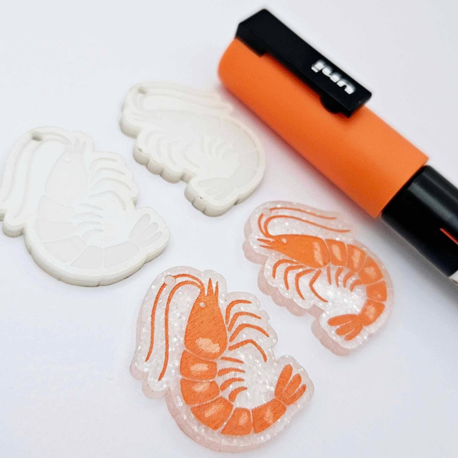 One pair of fairy dust glitter acrylic engraved prawn dangle earring blanks with protective paper on alongside a paint filled pair, laying flat with an orange posca paint pen