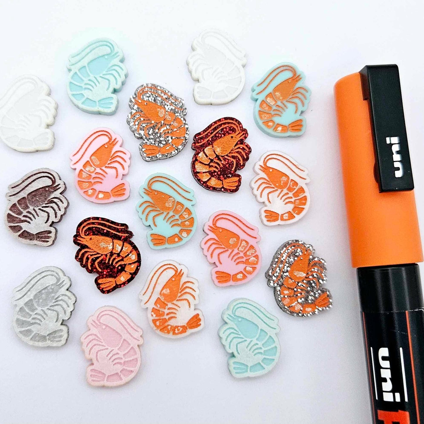 A selection of paint-filled prawn stud earring blanks in Pastel Pink, Pastel Aqua, Red Glitter, Silver Glitter and White Marble acrylic, with multiple unpainted pieces still covered with protective paper, pictured alongside an Orange Posca Paint Pen