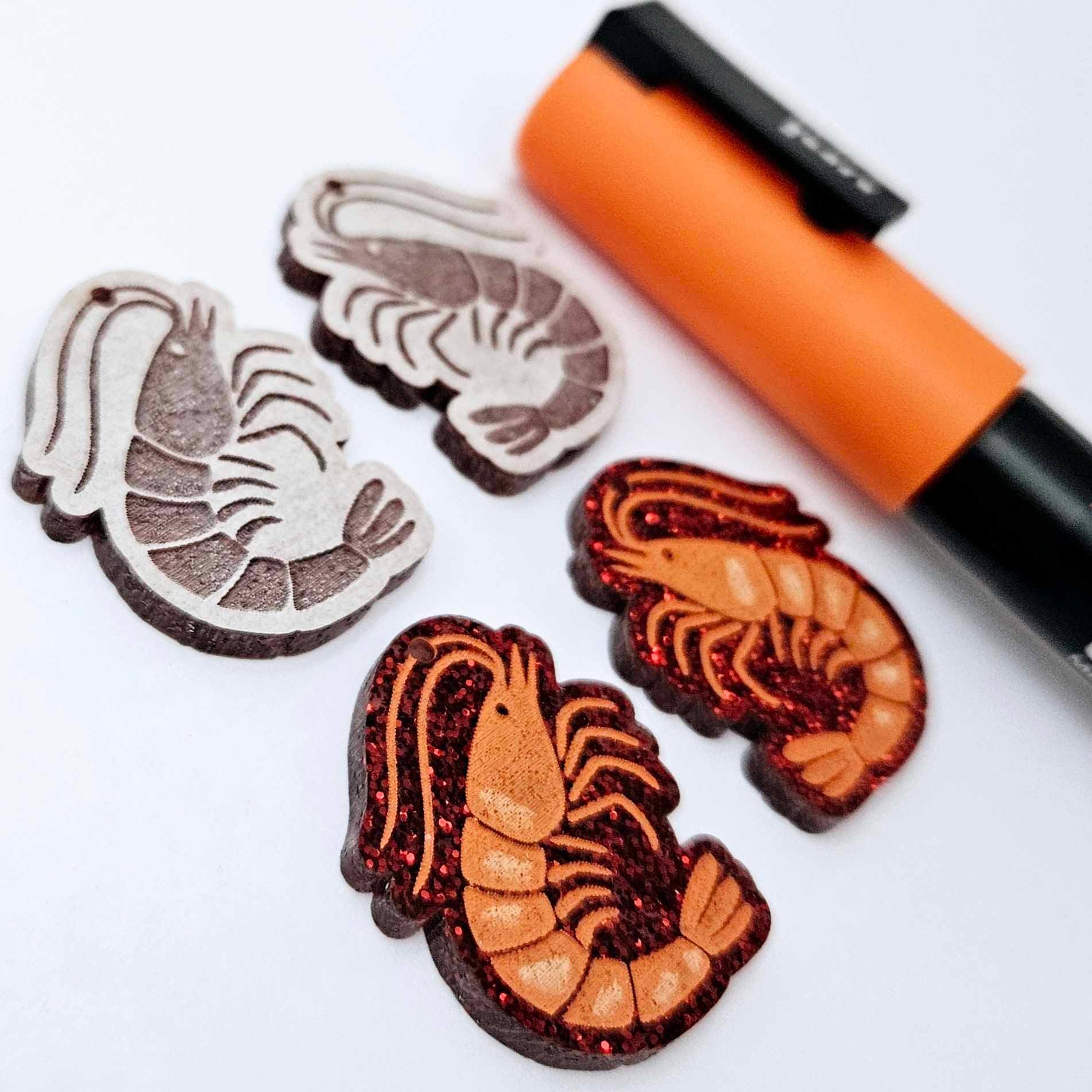 One pair of red glitter acrylic engraved prawn dangle earring blanks with protective paper on alongside a paint filled pair, laying flat with an orange posca paint pen