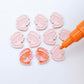 One paint-filled pair of pastel pink acrylic prawn stud earring blanks with multiple unpainted pieces still covered with protective paper, pictured with an uncapped Orange Posca Paint Pen ready for paint filling