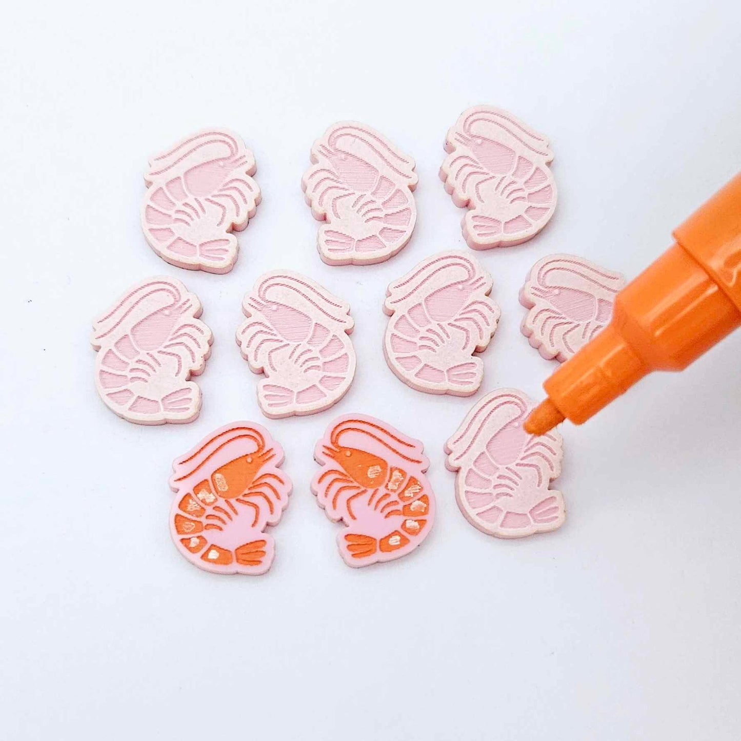 One paint-filled pair of pastel pink acrylic prawn stud earring blanks with multiple unpainted pieces still covered with protective paper, pictured with an uncapped Orange Posca Paint Pen ready for paint filling