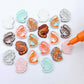 A selection of paint-filled prawn stud earring blanks in Pastel Pink, Pastel Aqua, Red Glitter, Silver Glitter and White Marble acrylic , with multiple unpainted pieces still covered with protective paper, pictured with an uncapped Orange Posca Paint Pen ready for paint filling