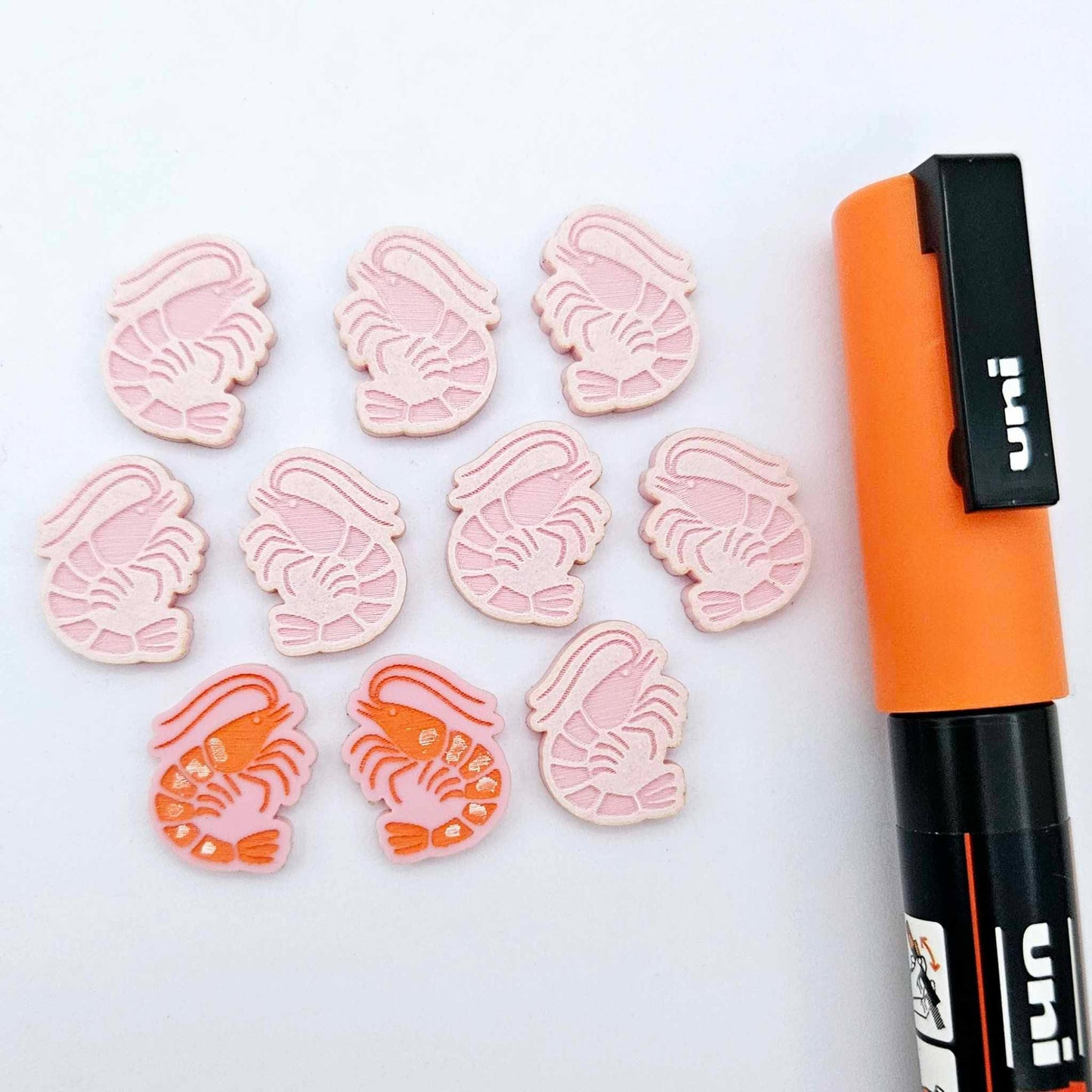 One paint-filled pair of pastel pink acrylic prawn stud earring blanks with multiple unpainted pieces still covered with protective paper, pictured alongside an Orange Posca Paint Pen