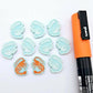 One paint-filled pair of pastel aqua acrylic prawn stud earring blanks with multiple unpainted pieces still covered with protective paper, pictured alongside an Orange Posca Paint Pen