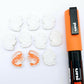 One paint-filled pair of White Marble acrylic prawn stud earring blanks with multiple unpainted pieces still covered with protective paper, pictured alongside an Orange Posca Paint Pen