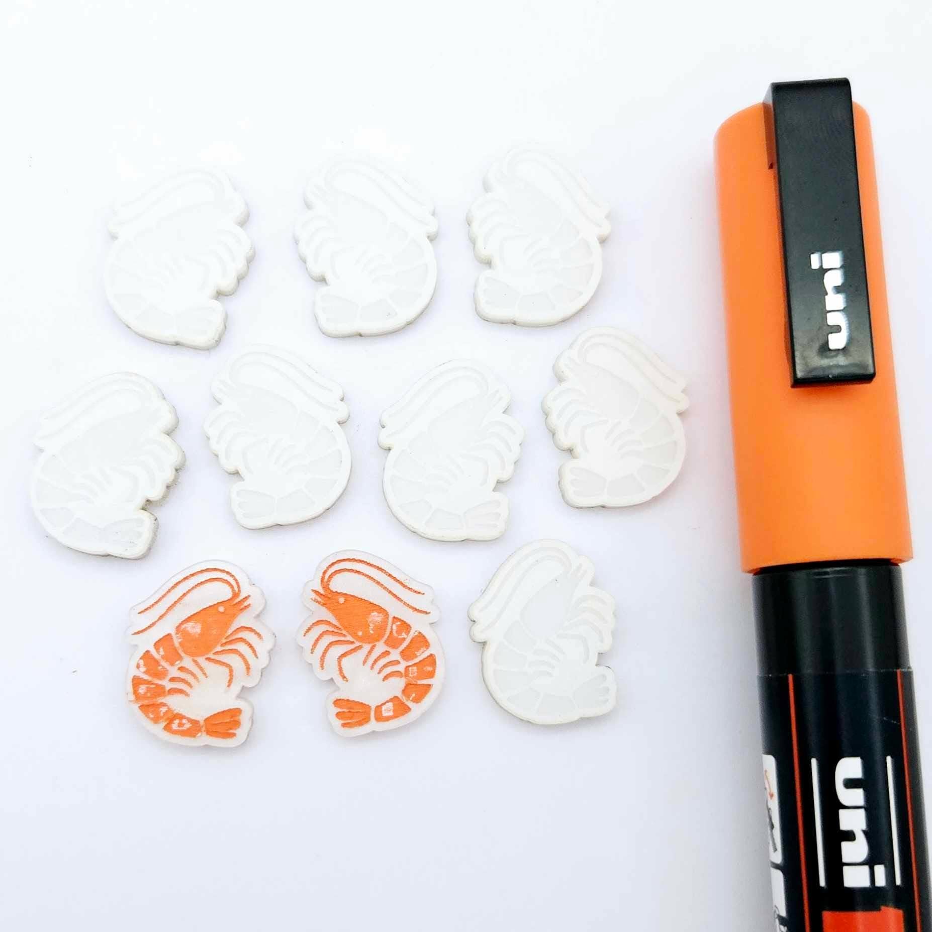One paint-filled pair of White Marble acrylic prawn stud earring blanks with multiple unpainted pieces still covered with protective paper, pictured alongside an Orange Posca Paint Pen