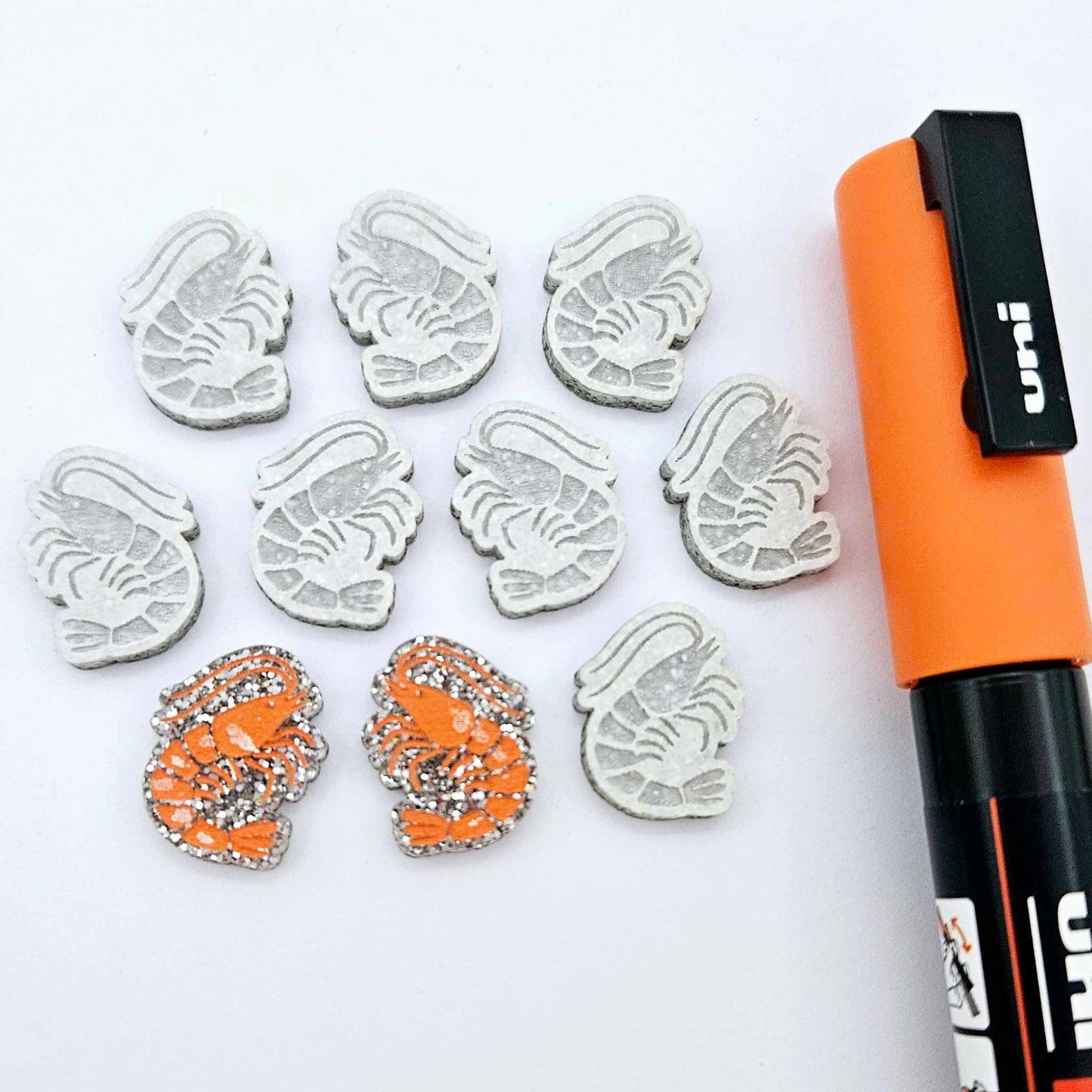 One paint-filled pair of silver glitter acrylic prawn stud earring blanks with multiple unpainted pieces still covered with protective paper, pictured alongside an Orange Posca Paint Pen