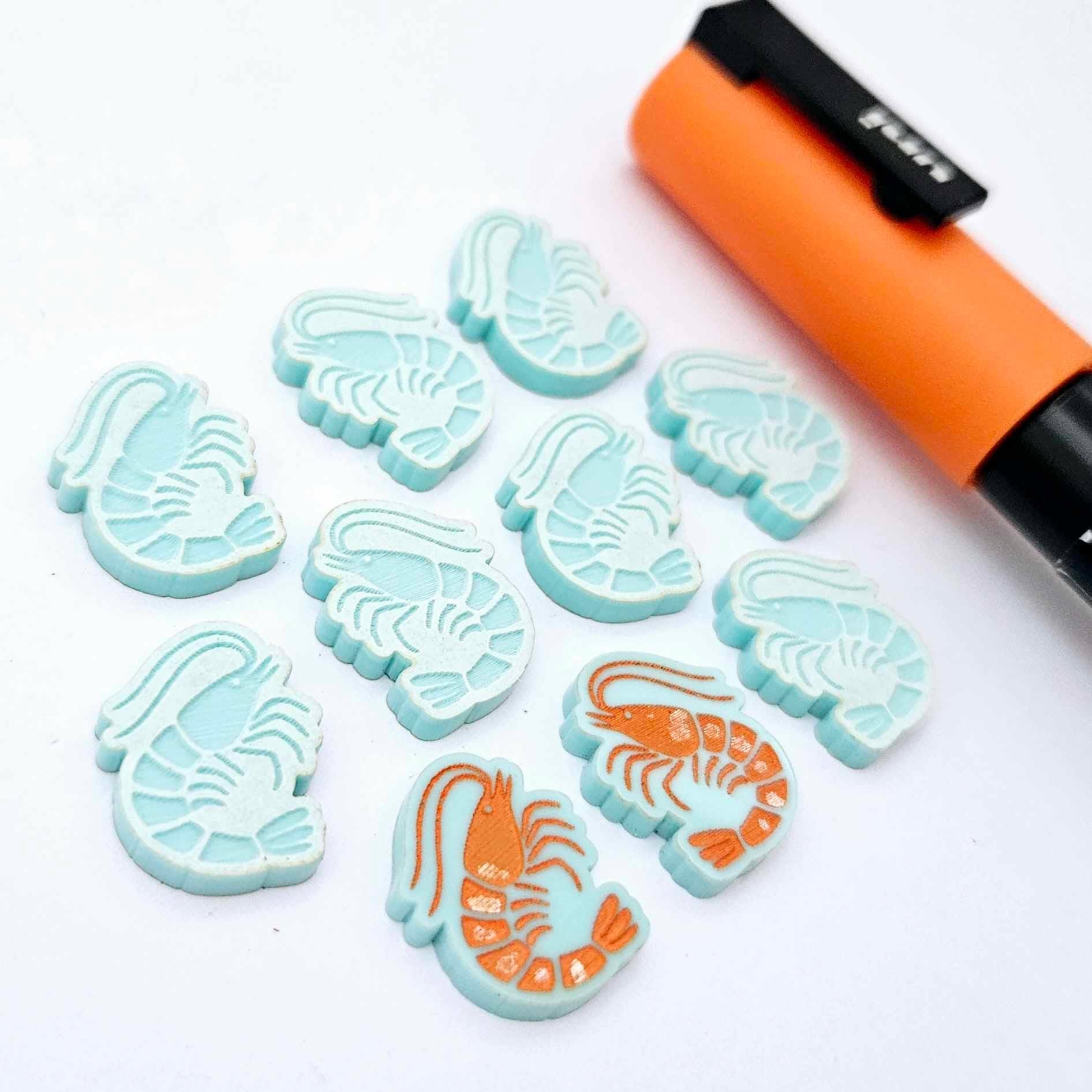 One paint-filled pair of pastel aqua acrylic prawn stud earring blanks with multiple unpainted pieces still covered with protective paper, pictured alongside an Orange Posca Paint Pen