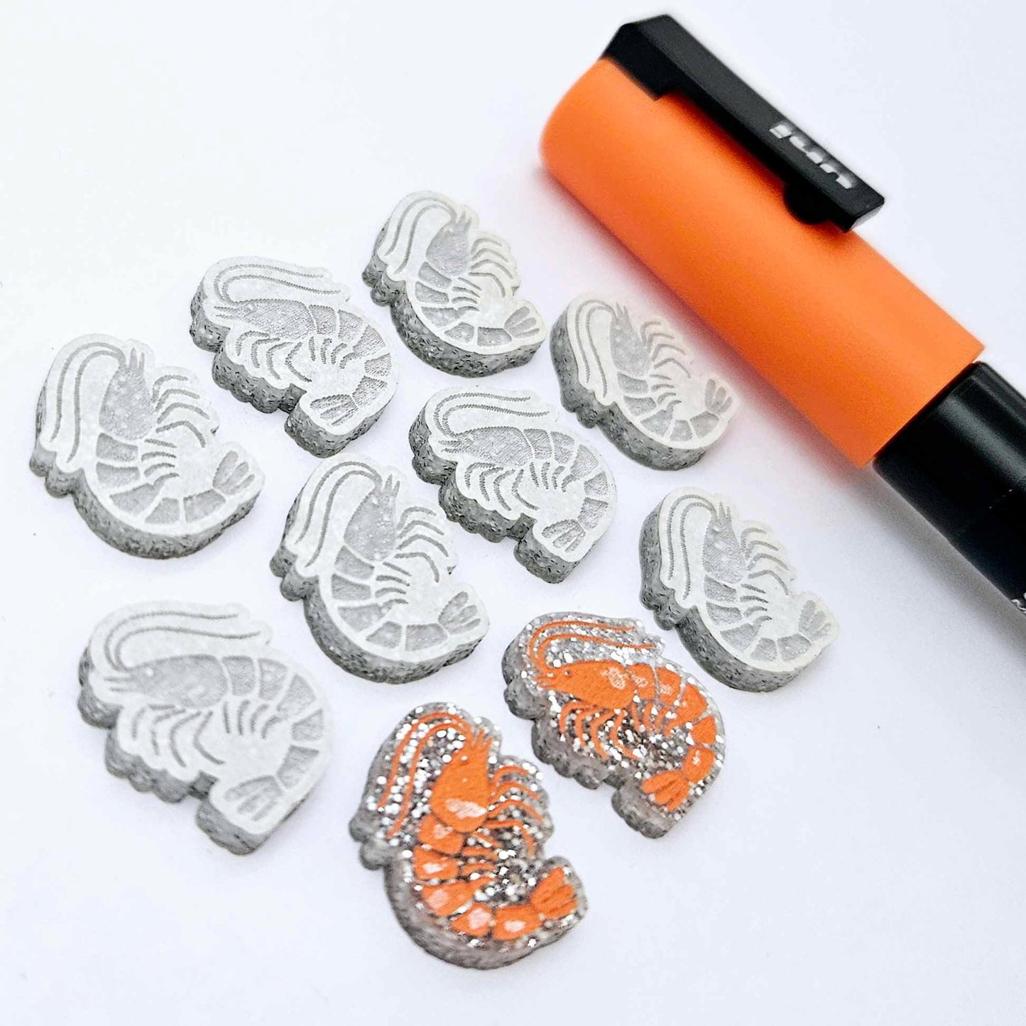 One paint-filled pair of silver glitter acrylic prawn stud earring blanks with multiple unpainted pieces still covered with protective paper, pictured alongside an Orange Posca Paint Pen