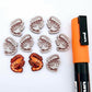 One paint-filled pair of red glitter acrylic prawn stud earring blanks with multiple unpainted pieces still covered with protective paper, pictured alongside an Orange Posca Paint Pen