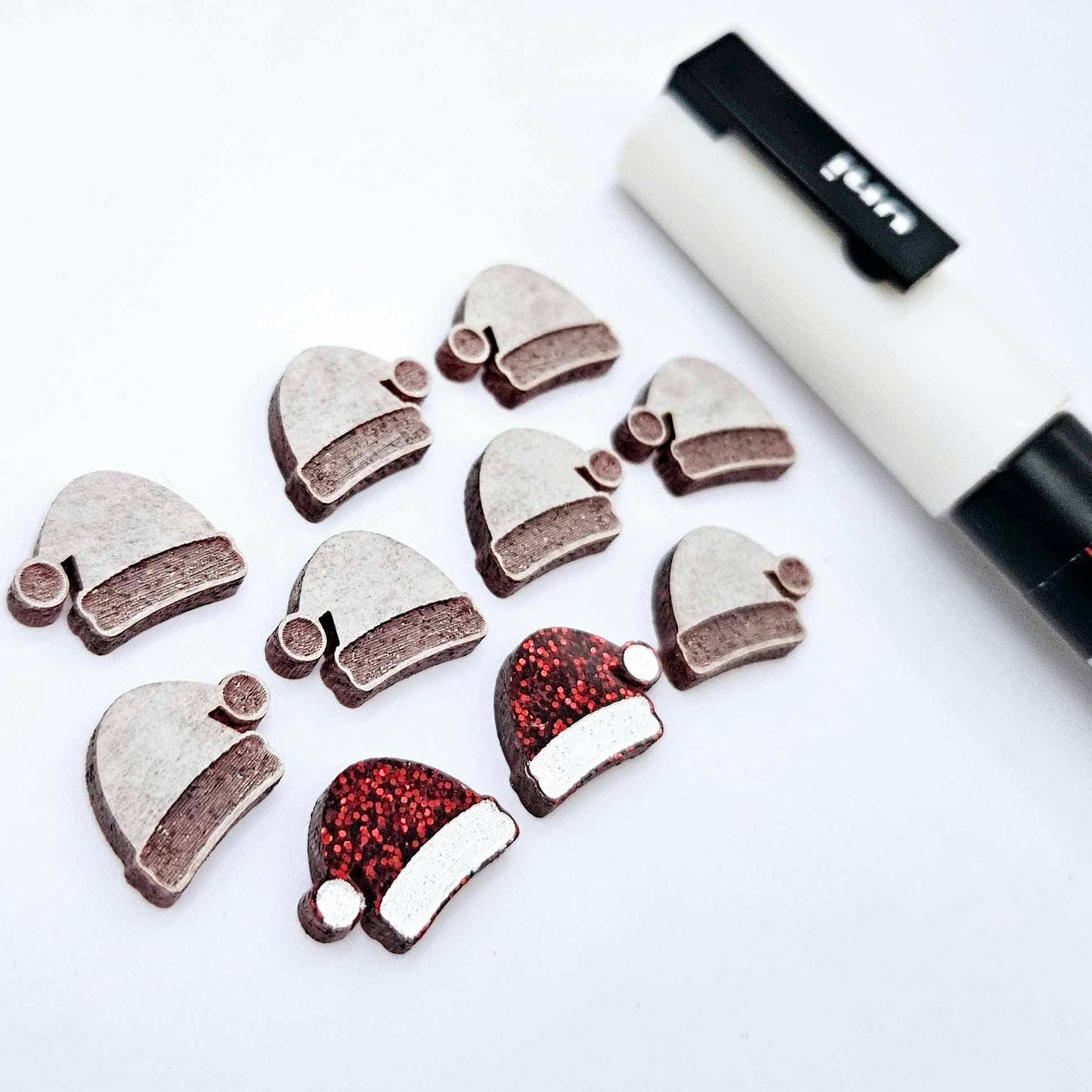 One paint-filled pair of red glitter acrylic Santa hat stud blanks alongside multiple unpainted pieces still covered with protective paper, pictured with a White Posca Paint Pen