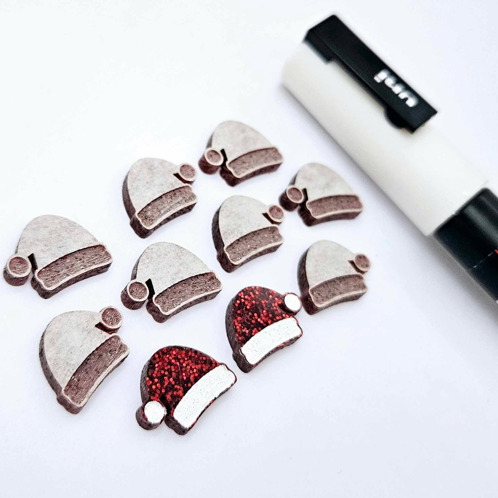 One paint-filled pair of red glitter acrylic Santa hat stud blanks alongside multiple unpainted pieces still covered with protective paper, pictured with a White Posca Paint Pen